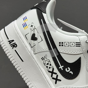 Peut inclure: Chaussure de sport blanche avec des d&eacute;tails noirs et le mot "AIR" imprim&eacute; sur le c&ocirc;t&eacute;. La chaussure pr&eacute;sente un logo noir en forme de virgule et divers motifs graphiques, dont un c&oelig;ur, un signe plus et le texte "CULT OF COMS".