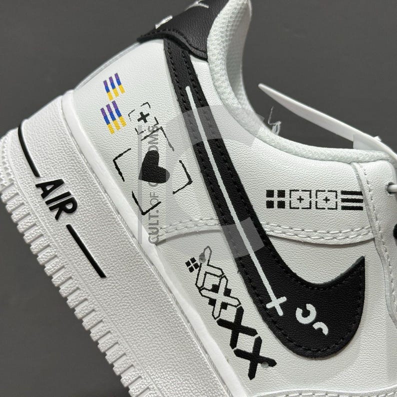 Peut inclure: Chaussure de sport blanche avec des d&eacute;tails noirs et le mot "AIR" imprim&eacute; sur le c&ocirc;t&eacute;. La chaussure pr&eacute;sente un logo noir en forme de virgule et divers motifs graphiques, dont un c&oelig;ur, un signe plus et le texte "CULT OF COMS".