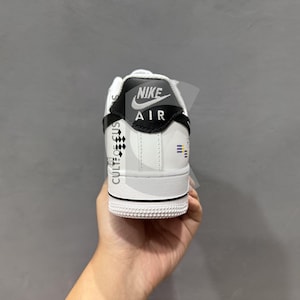 Peut inclure: Baskets Nike Air Force 1 blanches et noires. Le talon pr&eacute;sente le logo Nike Air en noir et blanc. Le c&ocirc;t&eacute; de la chaussure porte le texte "CULT OF US" et un petit motif &agrave; carreaux. L'autre c&ocirc;t&eacute; comporte trois rayures bleues et jaunes.