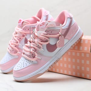 May include: Pair of pink and white trainers with a heart-shaped tag. The shoes have pink laces, a white swoosh, and a pink interior. They are displayed next to a matching pink box with a grid pattern.