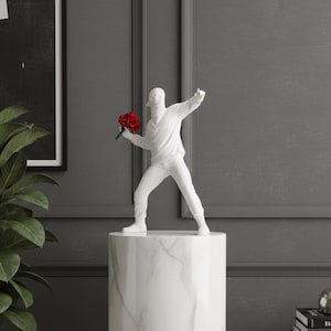 Peut inclure: Une sculpture blanche d'une figure lançant un bouquet de fleurs rouges, montée sur un piédestal en marbre. La sculpture est placée contre un mur gris avec une œuvre encadrée et une plante en pot.