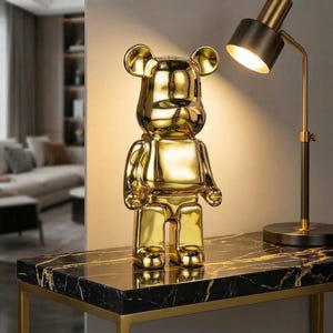Puede incluir: Una figura de oso dorada y estilizada con un acabado brillante. El oso tiene un diseño en bloques con orejas y extremidades redondeadas. Se encuentra sobre una mesa negra con vetas de mármol y un marco dorado, junto a una lámpara de escritorio de latón.
