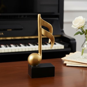 Peut inclure: Une sculpture dorée en forme de croche sur une base rectangulaire noire. La sculpture est posée sur une surface en bois devant un piano. Une rose blanche dans un vase en verre est en arrière-plan.