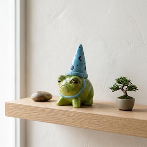 Peut inclure: Figurine de grenouille verte en céramique portant un chapeau de sorcier bleu orné d'étoiles blanches et de motifs floraux. La grenouille a une expression grincheuse et une mentonnière bleue. Un petit bonsaï et une pierre lisse sont également sur l'étagère en bois.