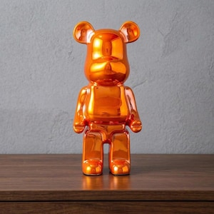 Può includere: Statuetta decorativa a forma di orso arancione lucido. L'orso ha un design squadrato con orecchie e arti arrotondati. È posizionato su una superficie di legno contro un muro grigio. L'oggetto è un pezzo decorativo vivace.