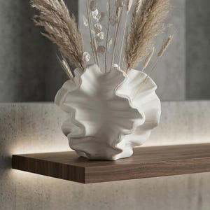 Peut inclure: Un vase en céramique blanc, froissé, contenant des herbes ornementales séchées et des gousses de graines. Le vase est posé sur une étagère en bois foncé avec un éclairage chaleureux. L'ensemble est contre un fond gris neutre.