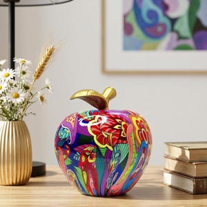 Puede incluir: Figura decorativa de manzana con un vibrante diseño floral multicolor. La manzana tiene un tallo y una hoja dorados. La figura está sobre una superficie de madera, con un jarrón dorado con flores y una pila de libros en el fondo.