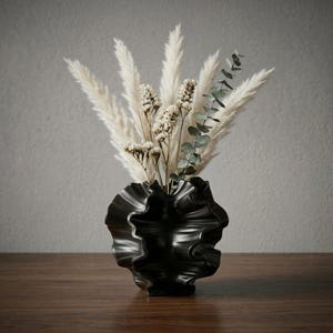 Peut inclure: Un vase en céramique noir au design froissé contient un bouquet d'herbe de la pampa séchée, de petites fleurs blanches et des branches d'eucalyptus. Le vase est posé sur une surface en bois foncé sur fond gris.