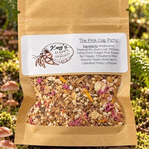 May include: A brown paper pouch containing a dry food mix labelled The Pink Cap Picnic by Kay's Happy Hermits. The label lists ingredients including mushrooms, mealworms, duck liver, minnows, oats, dragon fruit, and various seeds and petals.