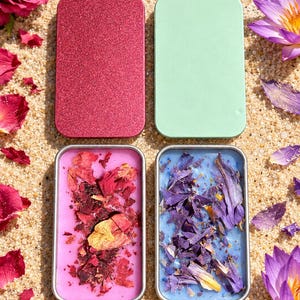 May include: Two rectangular metal tins with lids. One tin is filled with pink wax topped with dried red rose petals. The other tin contains blue wax topped with dried purple flower petals. The lids are glittery red and matte light green.