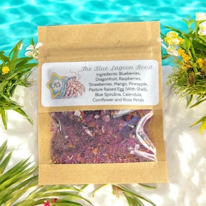 Peut inclure: Un petit sachet en papier brun avec une fenêtre transparente, contenant un mélange coloré d'ingrédients. Le sachet porte une étiquette indiquant "The Blue Lagoon Boost" et énumère des ingrédients comme les myrtilles, le fruit du dragon et les pétales de rose. L'arrière-plan est flou.