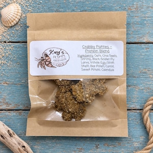 May include: A brown paper pouch with a clear window containing textured brown food patties. The label reads: Kay's Happy Hermits, Crabby Patties - Protein Blend. Ingredients: Oats, Chia Seeds, Shrimp, Black Soldier Fly Larva, Whole Egg, Bee Pollen, Carrot, Sweet Potato, Calendula.
