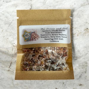 Peut inclure: Un petit sac en papier brun avec une fenêtre transparente contenant un mélange d'ingrédients. Une étiquette blanche affiche le texte "The Protein and Peel" et une liste d'ingrédients, dont de la mue de serpent, des supervers et des graines de chanvre. L'étiquette comporte également un logo.