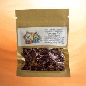 May include: A brown paper bag with a clear window, filled with a mix of dried ingredients. A white label displays the text "The Volcanic Bloom" and a list of ingredients, including dragonfruit and mealworms. The label also features a cartoon hermit crab.