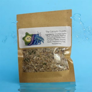 May include: A brown paper bag with a clear window showing a mix of ingredients. The bag features a white label with the text "The Calcium Crunch" and a list of ingredients, including crushed oyster shell and pumpkin seeds. A blue hermit crab logo is also present.