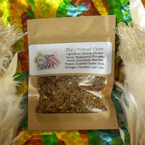 May include: A small brown paper bag with a clear window, containing a mix of ingredients. The label reads "The Primal Core" and lists ingredients including shrimp, chicken hearts, and pumpkin seeds. The background is colourful.