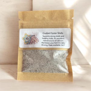 Peut inclure: Un sachet en papier brun contenant des coquilles d'huîtres broyées. L'étiquette blanche présente un bernard-l'ermite en dessin animé dans une étoile et le texte : Crushed Oyster Shells, Supports strong shells and healthy molts. An excellent natural source of calcium that keeps your hermit crabs thriving. Make available 24/7.