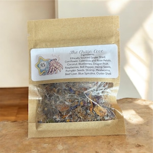 Peut inclure: Un petit sachet transparent rempli d'un mélange d'ingrédients séchés, dont des graines et des pétales, à l'intérieur d'une pochette en papier brun. L'étiquette indique "The Chitin Cove" avec une liste d'ingrédients. Un logo avec un crabe est sur l'étiquette.