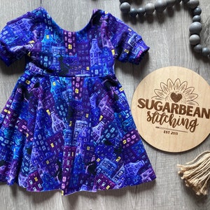May include: A purple and blue dress with a city skyline print. The dress has short sleeves and a round neckline. The dress is on a wooden surface with a sign that says "Sugarbean Stitching Est 2011".