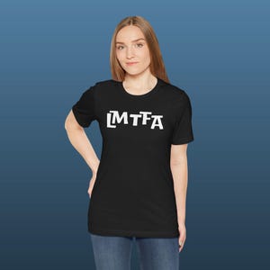 Puede incluir: Camiseta negra con el texto blanco "MTFA" en una fuente en negrita y cuadrada. La camiseta es de cuello redondo y parece estar hecha de un material suave. El fondo es un degradado de azul.
