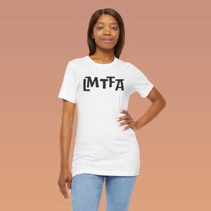 Puede incluir: Una camiseta blanca con el texto negro "MTFA" impreso en la parte delantera. La modelo lleva vaqueros azules. El fondo es de color naranja apagado.