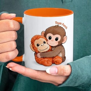 May include: White ceramic mug with an orange interior and handle. The mug features a cartoon illustration of two hugging monkeys, with the text "Team Punch" in orange. The monkeys are brown and orange.
