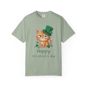 Op de afbeelding: Saliegroen T-shirt met een cartoonkat met een groene leprechaunhoed en klavers. De kat heeft een humeurige uitdrukking. De woorden "Happy St. Catrick's day" staan eronder in het groen.