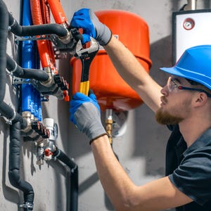 May include: A plumber wearing a blue hard hat and safety glasses uses a wrench on a network of pipes. The pipes are black, blue, and orange, and are connected to a red tank and other equipment. The setting appears to be a utility room.