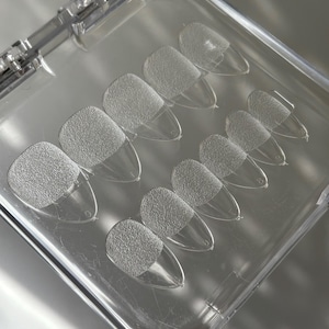May include: Clear, textured press-on nails in a clear plastic case. The nails have a textured, matte finish on the top half and a clear, pointed tip. The case is open, revealing the arrangement of the nails.
