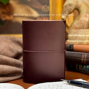 Könnte beinhalten: Ein dunkelbraunes Leder-Notizbuch, das mit einem dünnen Gummiband verschlossen ist. Der Einband hat eine glatte Textur und eine abgerundete obere Ecke. Über dem Notizbuch steht der Text: Made to be your companion for every chapter. Ein Füllfederhalter liegt daneben.