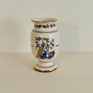Op de afbeelding: Witte keramische vaas met een voet, versierd met gouden randen en een pauwenontwerp in blauw en goud. De vaas heeft een uitlopende rand en een cilindrische body, geschikt voor bloemstukken.