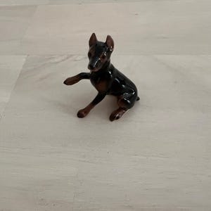 May include: A small, ceramic Doberman Pinscher figurine. The dog is black and brown with a glossy finish. It is posed with one paw raised, giving the impression of movement. The figurine is a decorative item.
