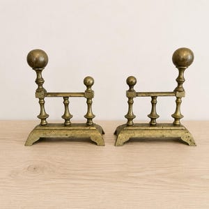 May include: A pair of matching brass andirons featuring a horizontal bar supported by three vertical posts, with a large spherical finial on the taller front post and a smaller spherical finial on the shorter rear post, set on a flat base.