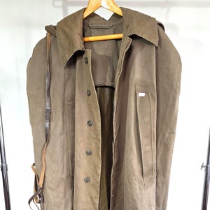 May include: A long, olive-drab trench coat featuring a wide collar, a front button closure, and a thin leather strap hanging from the side. The coat is constructed from a heavy, matte fabric and includes a vertical pocket flap on the chest area.