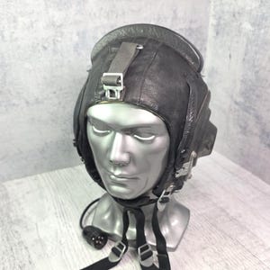 Può includere: Un casco da volo in pelle nera con padiglioni auricolari integrati, sottogola regolabili con fibbie in metallo e un cavo di comunicazione con connettore multipolare. Il casco è esposto su una testa di manichino color argento.