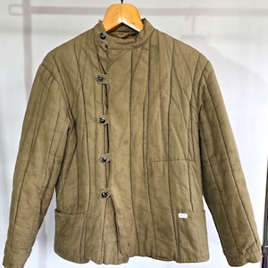 May include: A tan quilted jacket featuring a high collar, vertical stitching, and a side-fastening front closure with four dark buttons. The jacket includes two front pockets and a small white rectangular tag on the lower left pocket area.