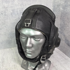Puede incluir: Un casco de aviador de cuero negro con orejeras integradas, una correa central ajustable con hebilla de metal y una correa para la barbilla con cierre metálico. El casco está diseñado con paneles laterales reforzados y costuras visibles en toda la estructura.