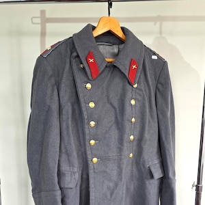 May include: A double-breasted charcoal gray wool overcoat featuring a wide collar with bright red tabs, each embroidered with a white cross. The coat includes two rows of gold-toned metal buttons, two side pockets with flaps, and shoulder epaulets.