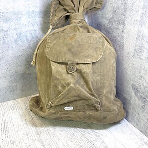 Op de afbeelding: Een beige canvas tas met trekkoordsluiting, voorzien van een voorvak met klep en een enkele knoopsluiting, en een klein wit rechthoekig label met de zwarte tekst 000352 aan de onderkant. De tas heeft een geplooide bovenkant met een trekkoord.