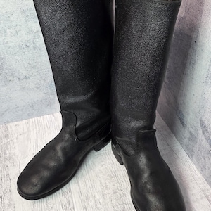 Puede incluir: Un par de botas de cuero negro con una superficie texturizada. Las botas tienen una ca&ntilde;a alta y una punta redondeada. Las botas est&aacute;n sobre una superficie de madera clara.