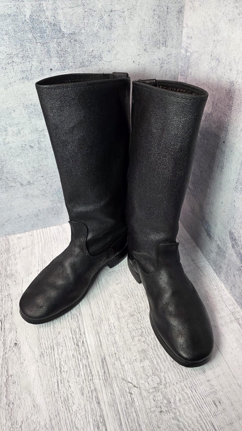 Puede incluir: Un par de botas de cuero negro con una superficie texturizada. Las botas tienen una ca&ntilde;a alta y una punta redondeada. Las botas est&aacute;n sobre una superficie de madera clara.