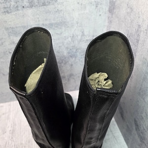 Puede incluir: Un par de botas de cuero negro con ca&ntilde;a alta. Las botas tienen un acabado liso y mate y un dise&ntilde;o cl&aacute;sico. El interior de cada bota contiene papel arrugado para soporte. Las botas est&aacute;n sobre un fondo neutro.