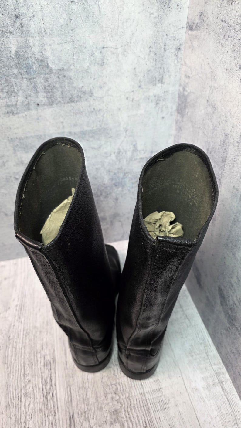 Puede incluir: Un par de botas de cuero negro con ca&ntilde;a alta. Las botas tienen un acabado liso y mate y un dise&ntilde;o cl&aacute;sico. El interior de cada bota contiene papel arrugado para soporte. Las botas est&aacute;n sobre un fondo neutro.