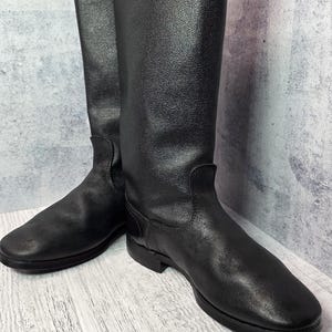 Puede incluir: Un par de botas de montar de cuero negro. Las botas tienen una ca&ntilde;a alta, una puntera redondeada y un tac&oacute;n bajo. El cuero tiene un aspecto texturizado. Las botas se exhiben sobre una superficie de madera clara.
