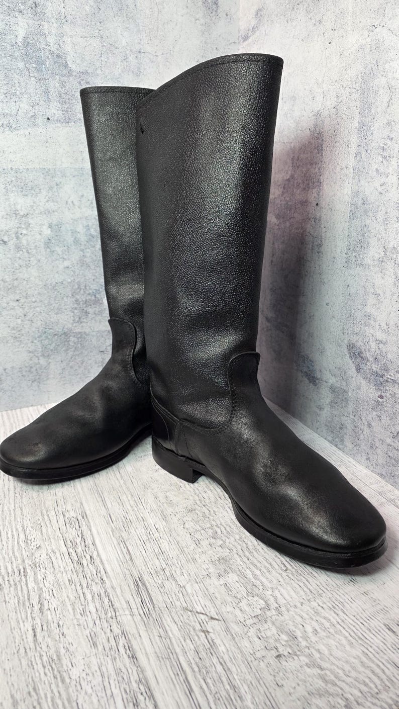 Puede incluir: Un par de botas de montar de cuero negro. Las botas tienen una ca&ntilde;a alta, una puntera redondeada y un tac&oacute;n bajo. El cuero tiene un aspecto texturizado. Las botas se exhiben sobre una superficie de madera clara.