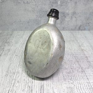 Puede incluir: Una cantimplora de metal de forma ovalada con un acabado plateado desgastado y un tapón de rosca de plástico negro. La superficie de la cantimplora muestra signos de uso, incluyendo pequeños arañazos e imperfecciones en el metal.
