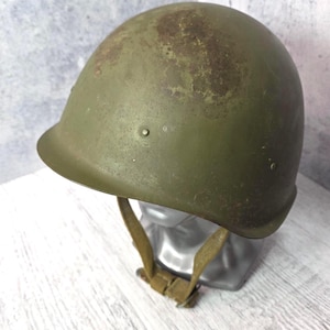 Può includere: Un elmetto militare verde oliva dall'aspetto usurato. L'elmetto ha una calotta arrotondata, una tesa e un sottogola. È esposto su una testa di manichino argentata.