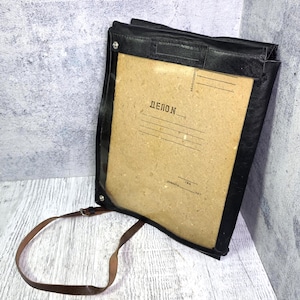 May include: Black leather document holder with a clear front, featuring the word "ДЕЛОN" printed on the interior paper. Includes a brown leather strap for carrying. The holder is on a white and gray surface.