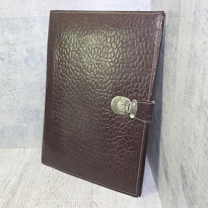 May include: A dark brown rectangular portfolio folder featuring a textured, pebble-like surface pattern. The folder is secured with a silver-toned metal latch mechanism attached to a small tab on the right side, with visible stitching along the edges.