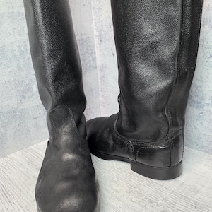 Puede incluir: Un par de botas negras de cuero hasta la rodilla. Las botas tienen un acabado liso y mate y una puntera redondeada. Las suelas son planas y las botas parecen sin usar. Las botas est&aacute;n de pie sobre un fondo gris claro.